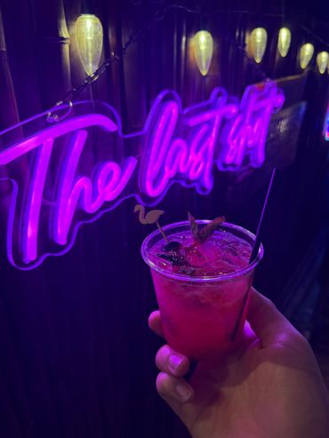 A hand holding a vibrant pink cocktail under purple neon lights.