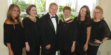 Group of six people dressed formally, smiling indoors by a window.