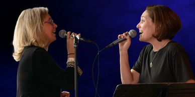 Two women passionately singing into microphones against a dark blue backdrop.