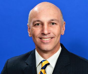 Smiling bald man in a suit with a blue and yellow tie against a blue background.