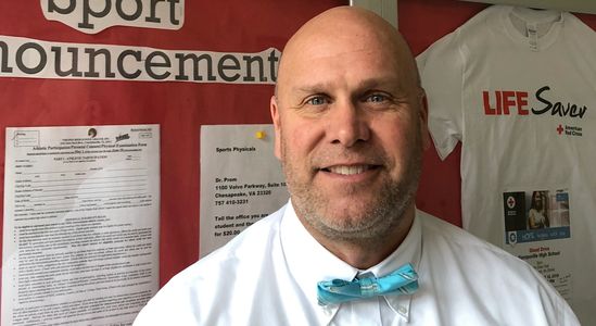 Smiling man in a white shirt and blue bow tie stands in front of a school announcement board.
