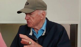 Elderly man in cap and jacket speaking with hand gestures.