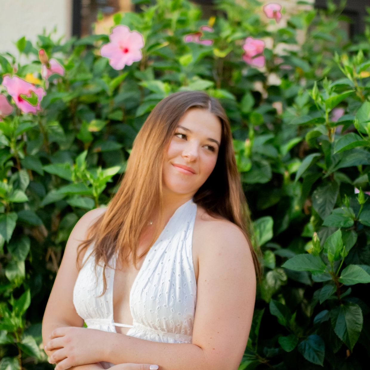 Young woman in white dress posing by pink flowers and green leaves.