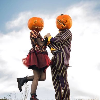 Couple wearing pumpkin heads holding hands outdoors.
