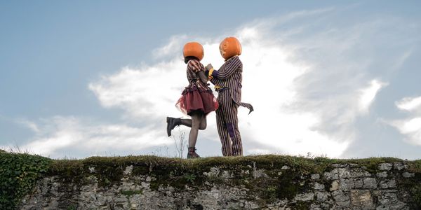 Two people with pumpkin heads holding hands on a stone wall.