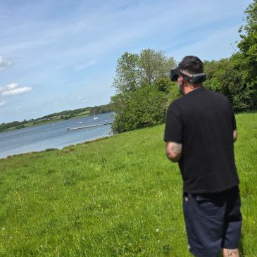 A man wearing a VR headset stands on a grassy field near a lake.