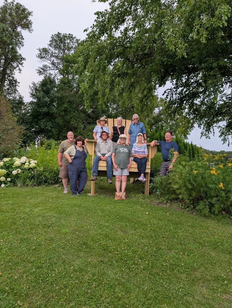 Group of people posing around a large wooden chair outdoors in a garden.