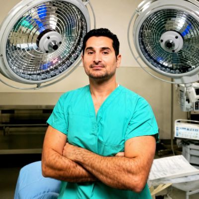Confident surgeon in green scrubs stands in operating room with surgical lights.