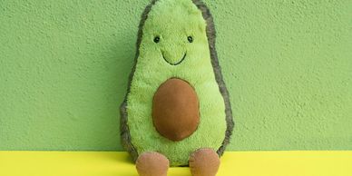 Cute avocado plush toy with a smile, sitting against a green wall.
