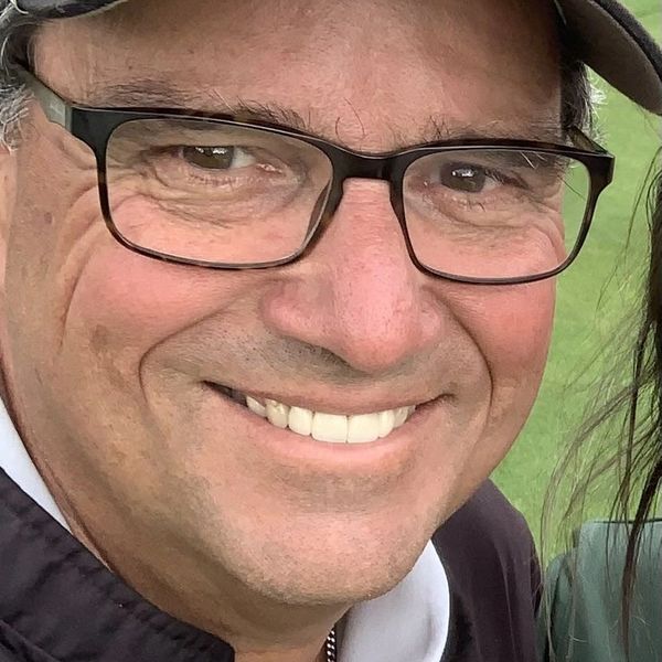 Smiling man wearing glasses and a Ptarmigan cap outdoors.