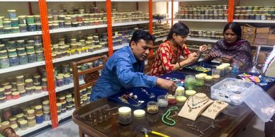 Three people crafting jewelry with beads in a workshop filled with jars of materials.