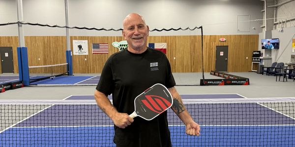 Smiling man holding a pickleball paddle on an indoor court.