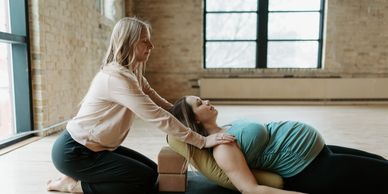 A yoga instructor assists a pregnant woman in a stretching pose indoors.