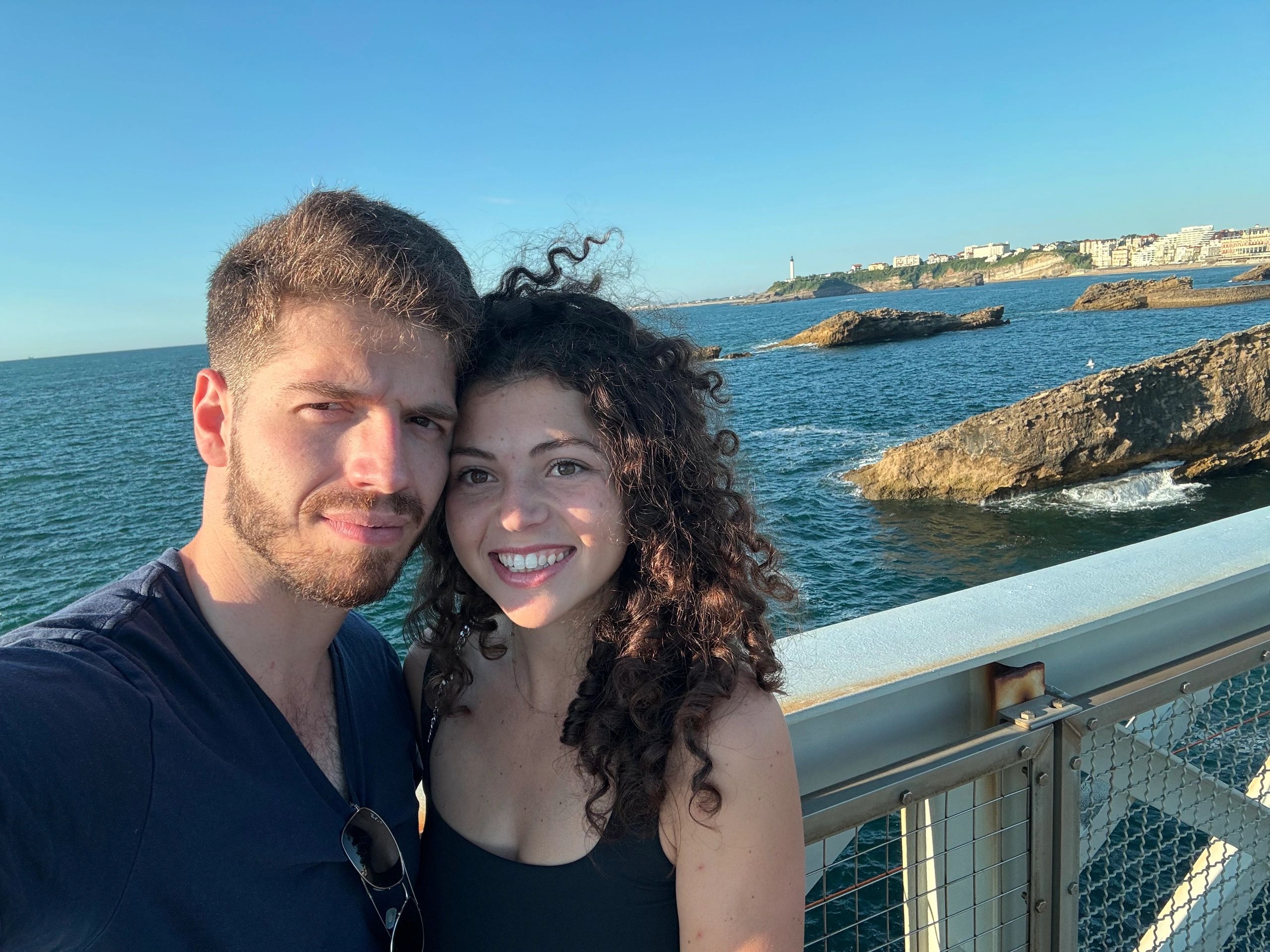A happy couple posing on a bridge with the ocean in the background.