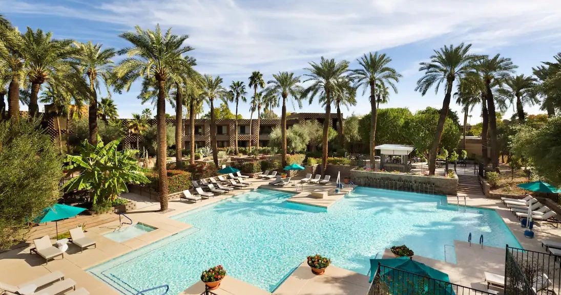 Resort pool surrounded by palm trees and lounge chairs under a blue sky.