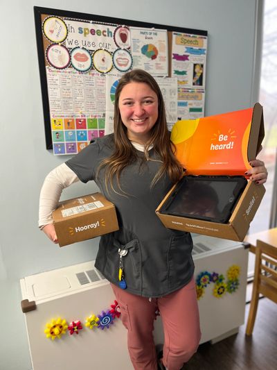 Young woman in scrubs holding two boxes with new AAC devices for clients