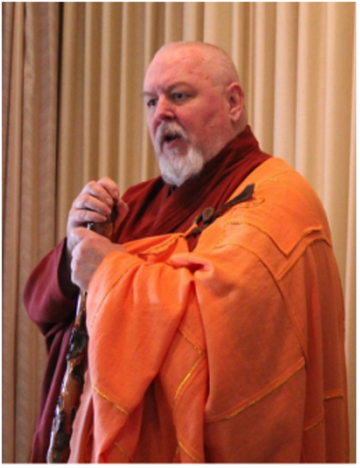 A heavyset man in his 60s with a shaved head and goatee, wearing red and orange monk's robes and hol