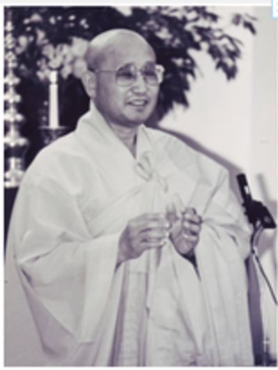 a black and white image of a Korean man with a shaved head wearing light-colored robes and large rou