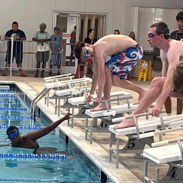 Swimmers preparing to dive as one athlete signals from the pool during a swim meet.