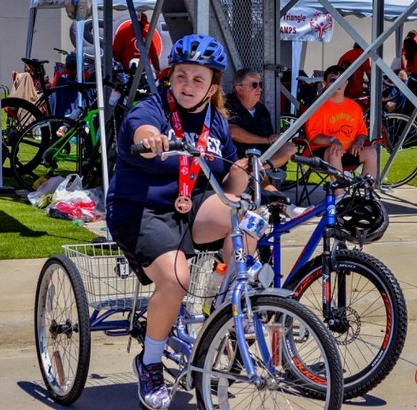 Young person rides a tricycle wearing a helmet and medal at an outdoor event.