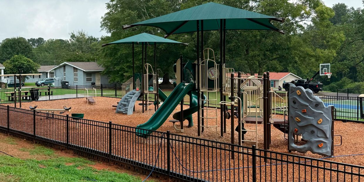 A fenced playground with slides, climbing wall, and shaded areas in a neighborhood.