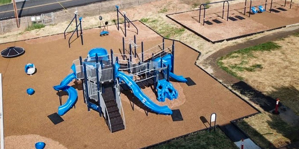 A playground with blue slides and swings on a brown rubber surface.