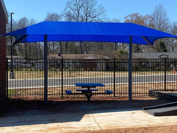 Blue shade canopy over a picnic table in an outdoor area with a metal fence backdrop.