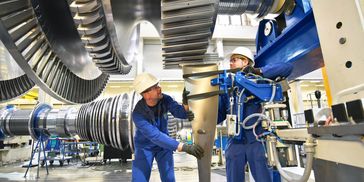 Two workers wearing blue uniforms and hard hats are in an industrial setting, examining large.