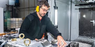 A man wearing safety goggles and yellow earmuffs operates a CNC machine in an industrial workshop.