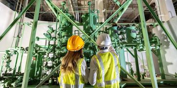 Two engineers in hard hats and high-visibility vests examine industrial machinery.