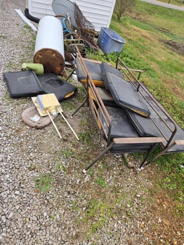 Old, rusty metal bed frame with torn cushions and various discarded items outside a building.