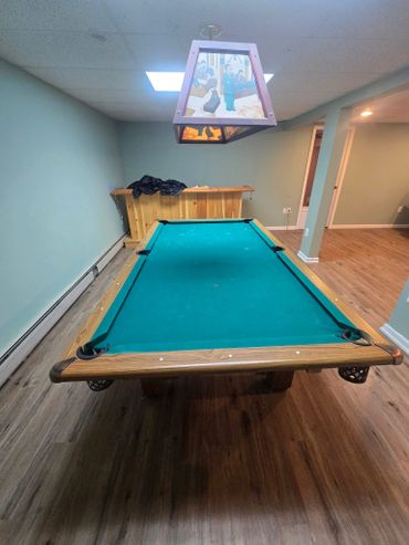 Empty pool table in a cozy basement game room with wooden bar and vintage light fixture.