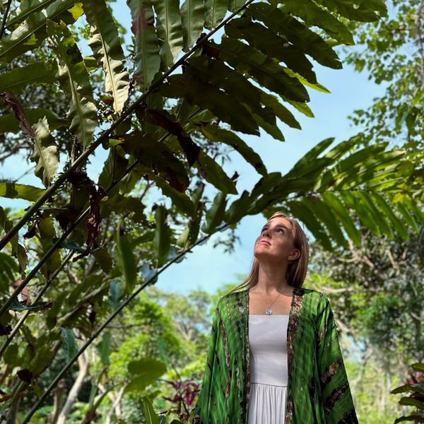 Woman in white dress and green shawl stands amid lush greenery looking up.