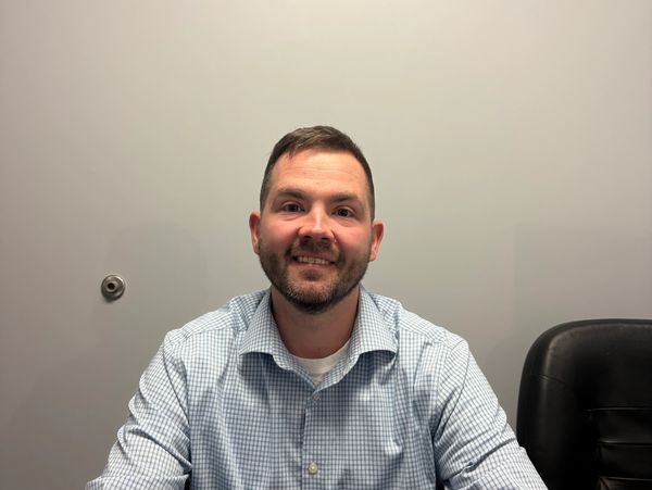 Man smiling, wearing a checkered shirt, seated against a plain wall.