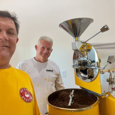Two men smiling near a yellow coffee roasting machine with beans in the cooling tray.