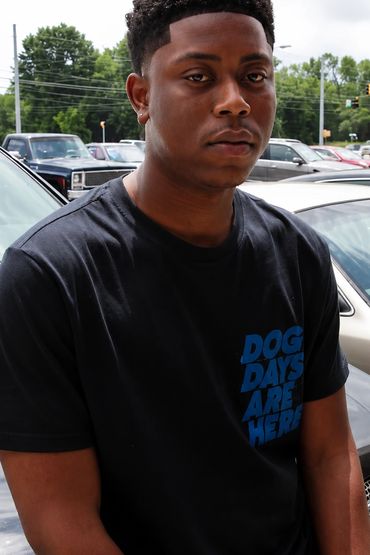 Young man wearing a black t-shirt with text, standing in a parking lot.