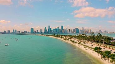 A coastal city skyline with turquoise waters and palm trees under a partly cloudy sky.