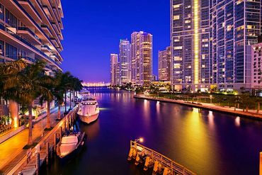 Night view of a waterfront city with illuminated skyscrapers and boats docked along the shore.