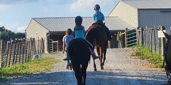 Two children riding horses on a gravel path near barns on a sunny day.