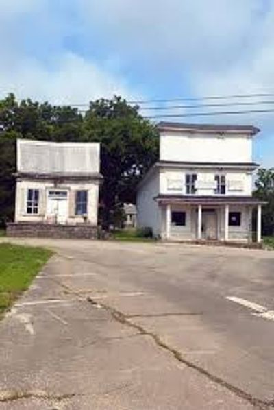 Two old small-town storefront buildings sit side-by-side along a cracked road, with trees.