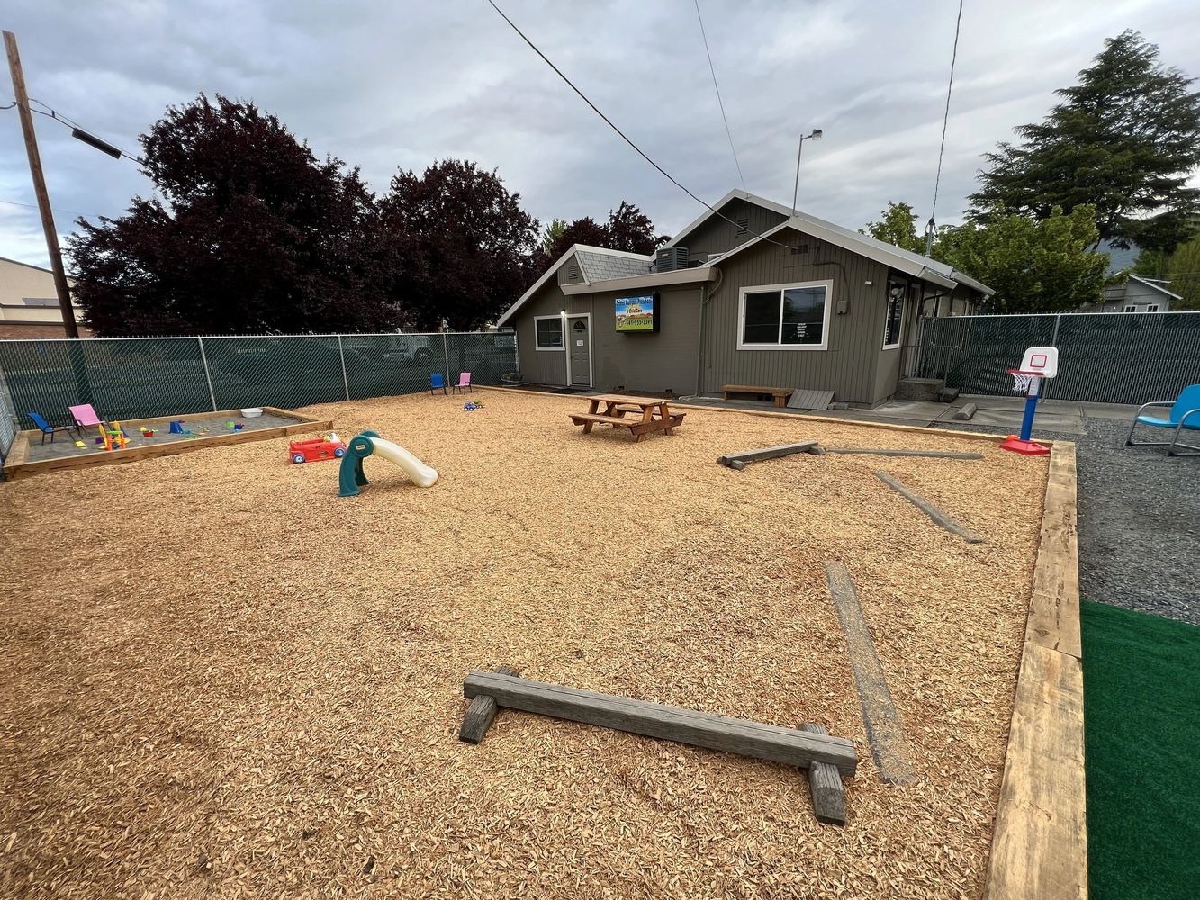 Outdoor children's play area with wood chips, small slide, sandbox, and basketball hoop.