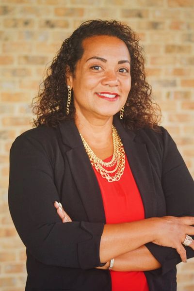 Confident woman in a red dress and black blazer stands with arms crossed.