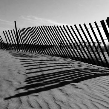 Black and white photo of a fence casting shadows on sand dunes.