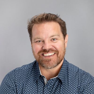 Smiling man with a beard wearing a patterned blue shirt against a grey background.