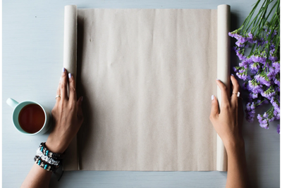 Hands rolling out blank brown paper with tea and purple flowers nearby.