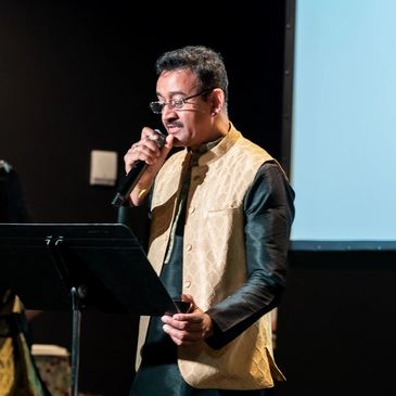 Man in traditional attire speaks into a microphone at a cultural event with an American flag nearby.