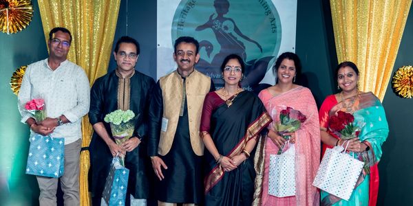 Six people dressed in traditional Indian attire holding flowers and gift bags at an event.
