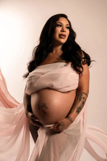 Pregnant woman draped in soft pink fabric with visible tattoos, looking away thoughtfully.