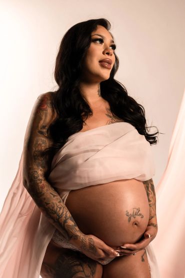 Pregnant woman with tattoos wrapped in soft fabric, looking hopeful.