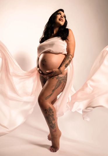 Pregnant woman with tattoos draped in flowing pink fabric poses gracefully.